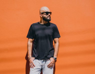 Turuncu bir duvarın arkasında siyah tişört giyen sakallı bir erkek model..