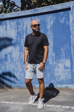 Mavi bir duvarın arkasında siyah, boş tişört giyen sakallı bir erkek model..