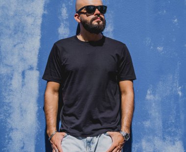 Mavi bir duvarın arkasında siyah, boş tişört giyen sakallı bir erkek model..