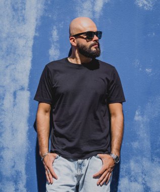 Mavi bir duvarın arkasında siyah, boş tişört giyen sakallı bir erkek model..