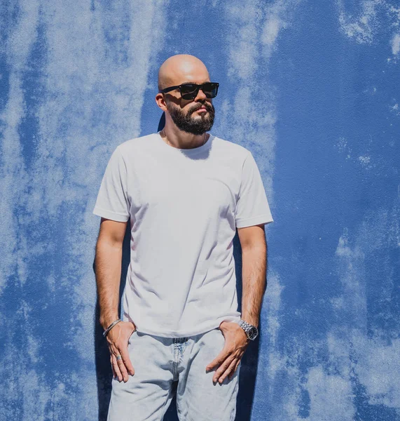 Mavi bir duvarın arkasında beyaz tişört giyen sakallı bir erkek model..