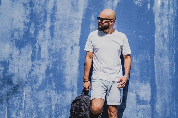Mavi bir duvarın arkasında beyaz tişört giyen sakallı bir erkek model..