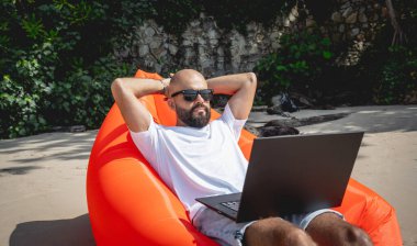 Genç adam şişme bir koltukta otururken kumsalda dizüstü bilgisayarıyla çalışıyor..