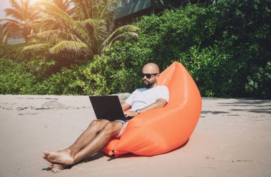 Genç adam şişme bir koltukta otururken kumsalda dizüstü bilgisayarıyla çalışıyor..