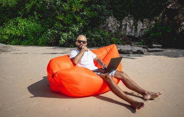 Genç adam şişme bir koltukta otururken kumsalda dizüstü bilgisayarıyla çalışıyor..