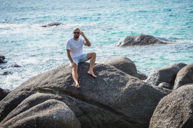 Genç adam kumsalda dinlenirken bir taşın üzerinde oturuyor..