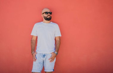 Turuncu bir duvarın arkasında beyaz tişört giyen sakallı bir erkek model..