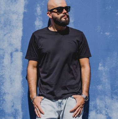 Mavi bir duvarın arkasında siyah, boş tişört giyen sakallı bir erkek model..