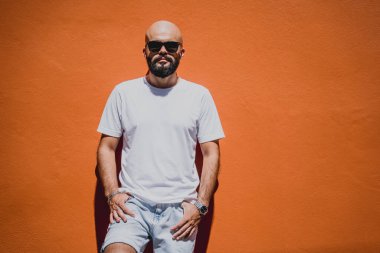 Turuncu bir duvarın arkasında beyaz tişört giyen sakallı bir erkek model..