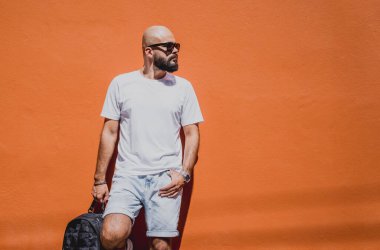 Turuncu bir duvarın arkasında beyaz tişört giyen sakallı bir erkek model..