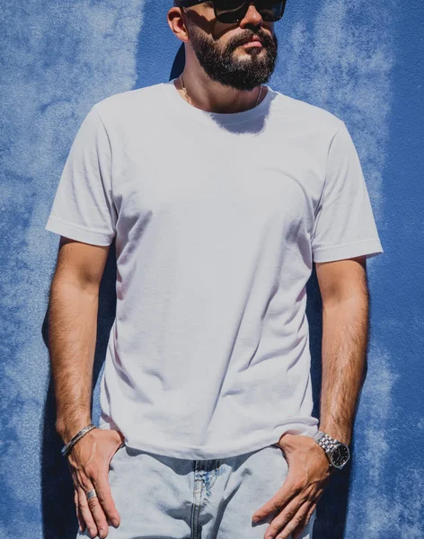 Male model with beard wearing white blank t-shirt on the background of an blue wall.