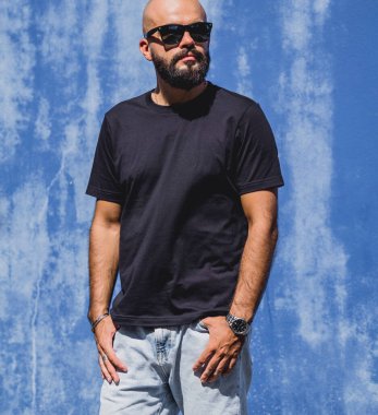 Mavi bir duvarın arkasında siyah, boş tişört giyen sakallı bir erkek model..