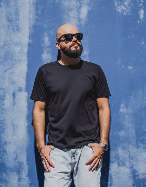 Mavi bir duvarın arkasında siyah, boş tişört giyen sakallı bir erkek model..