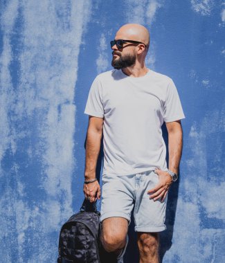 Mavi bir duvarın arkasında beyaz tişört giyen sakallı bir erkek model..