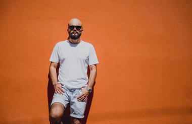 Turuncu bir duvarın arkasında beyaz tişört giyen sakallı bir erkek model..