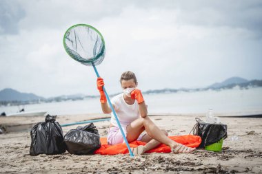 A female ecologist volunteer is resting after cleaning the beach on the seashore from plastic and other waste.