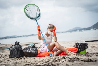 A female ecologist volunteer is resting after cleaning the beach on the seashore from plastic and other waste.