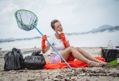 A female ecologist volunteer is resting after cleaning the beach on the seashore from plastic and other waste.