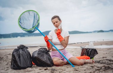 A female ecologist volunteer is resting after cleaning the beach on the seashore from plastic and other waste.