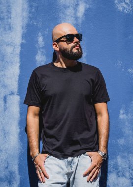 Mavi bir duvarın arkasında siyah, boş tişört giyen sakallı bir erkek model..