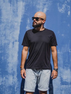 Mavi bir duvarın arkasında siyah, boş tişört giyen sakallı bir erkek model..