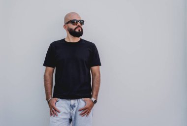 Beyaz bir duvarın arkasında siyah tişört giyen sakallı bir erkek model..