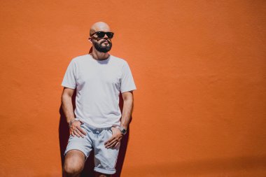 Turuncu bir duvarın arkasında beyaz tişört giyen sakallı bir erkek model..