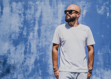 Mavi bir duvarın arkasında beyaz tişört giyen sakallı bir erkek model..