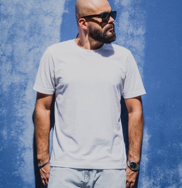 Mavi bir duvarın arkasında beyaz tişört giyen sakallı bir erkek model..