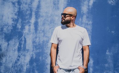 Mavi bir duvarın arkasında beyaz tişört giyen sakallı bir erkek model..