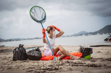 A female ecologist volunteer is resting after cleaning the beach on the seashore from plastic and other waste.
