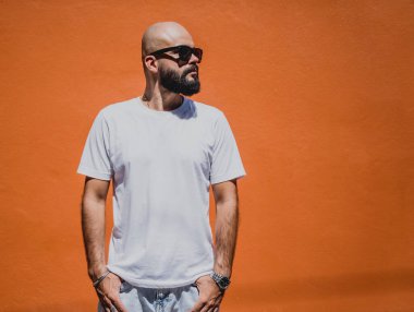 Turuncu bir duvarın arkasında beyaz tişört giyen sakallı bir erkek model..