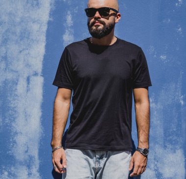 Mavi bir duvarın arkasında siyah, boş tişört giyen sakallı bir erkek model..