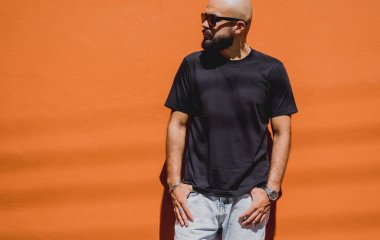 Turuncu bir duvarın arkasında siyah tişört giyen sakallı bir erkek model..