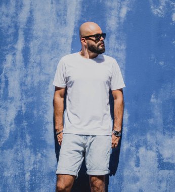 Mavi bir duvarın arkasında beyaz tişört giyen sakallı bir erkek model..