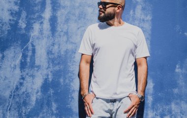 Mavi bir duvarın arkasında beyaz tişört giyen sakallı bir erkek model..