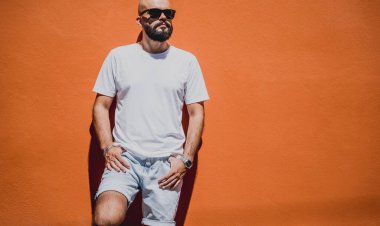 Turuncu bir duvarın arkasında beyaz tişört giyen sakallı bir erkek model..