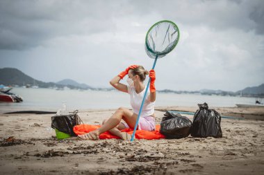 A female ecologist volunteer is resting after cleaning the beach on the seashore from plastic and other waste.