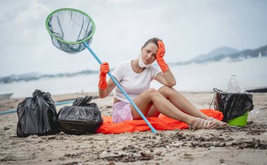 A female ecologist volunteer is resting after cleaning the beach on the seashore from plastic and other waste.