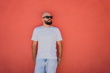 Turuncu bir duvarın arkasında beyaz tişört giyen sakallı bir erkek model..