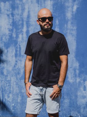 Mavi bir duvarın arkasında siyah, boş tişört giyen sakallı bir erkek model..