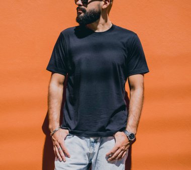 Turuncu bir duvarın arkasında siyah tişört giyen sakallı bir erkek model..