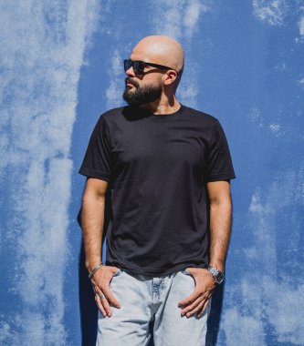 Mavi bir duvarın arkasında siyah, boş tişört giyen sakallı bir erkek model..