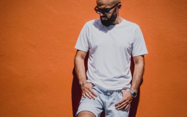 Turuncu bir duvarın arkasında beyaz tişört giyen sakallı bir erkek model..