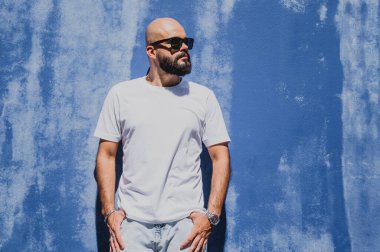 Mavi bir duvarın arkasında beyaz tişört giyen sakallı bir erkek model..