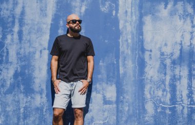 Mavi bir duvarın arkasında siyah, boş tişört giyen sakallı bir erkek model..