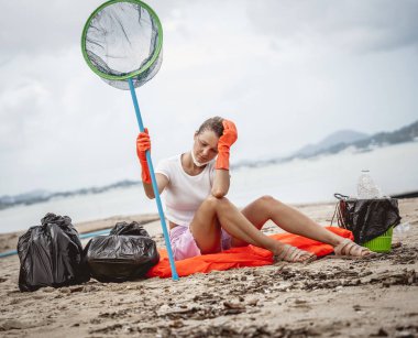 A female ecologist volunteer is resting after cleaning the beach on the seashore from plastic and other waste.