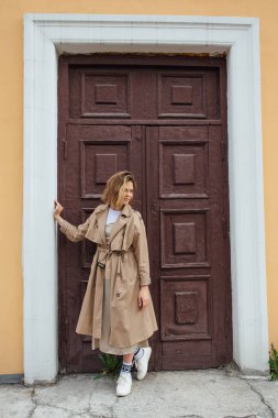 Genç, gülümseyen, vahşi saçlı, sonbahar montu giymiş, eski bir binanın kapısında poz veren bir kadın..