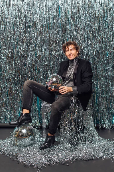 Uzun boylu çekici bir adam, ışıltılı arka planda yerde oturuyor ve ayna disko topu elinde şampanya kadehi tutuyor. Parti konsepti.