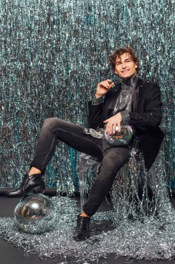 Uzun boylu çekici bir adam, ışıltılı arka planda yerde oturuyor ve ayna disko topu elinde şampanya kadehi tutuyor. Parti konsepti.